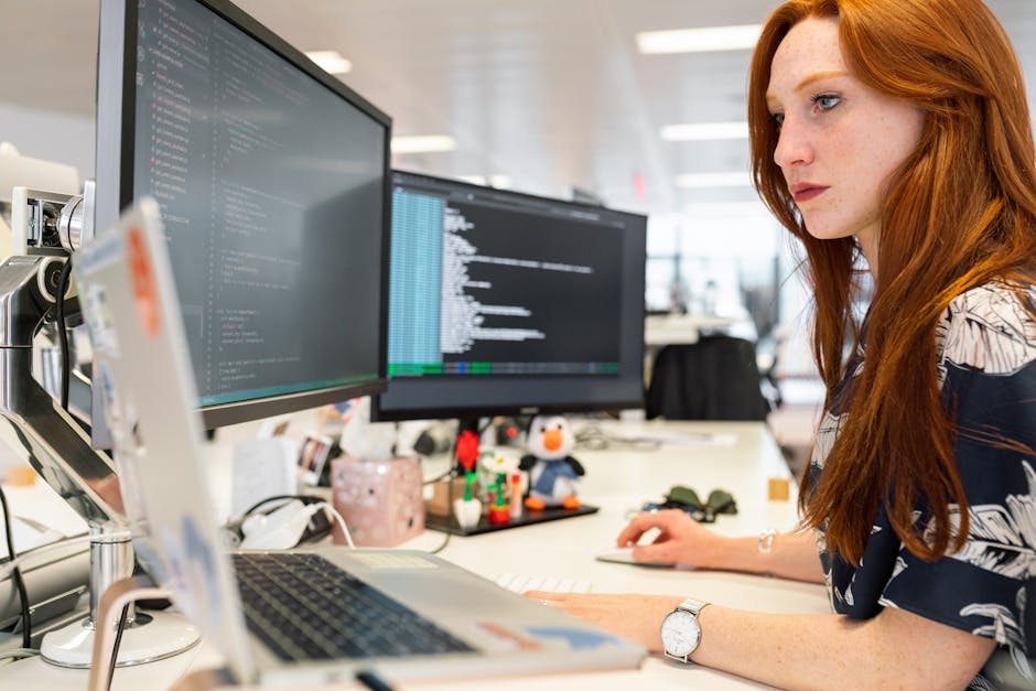 A focused female software engineer coding on dual monitors in a modern office