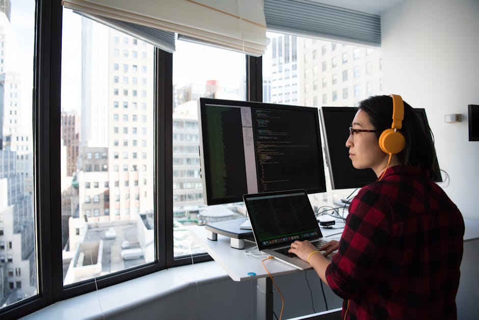 A businesswoman coding at a standing desk with city views through large windows, wearing headphones